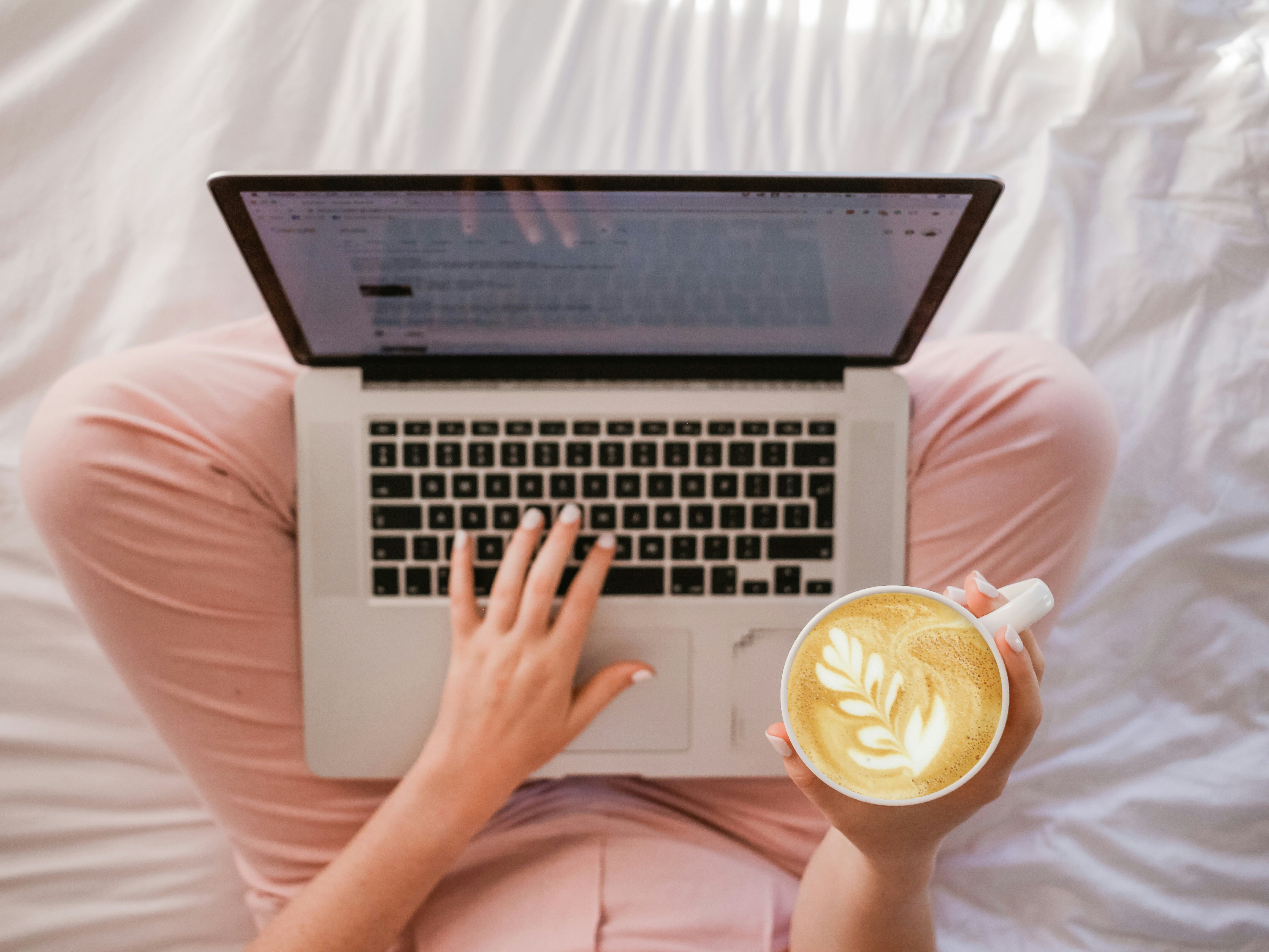 female typing on mac book sitting criss cross with cappuccino in hand